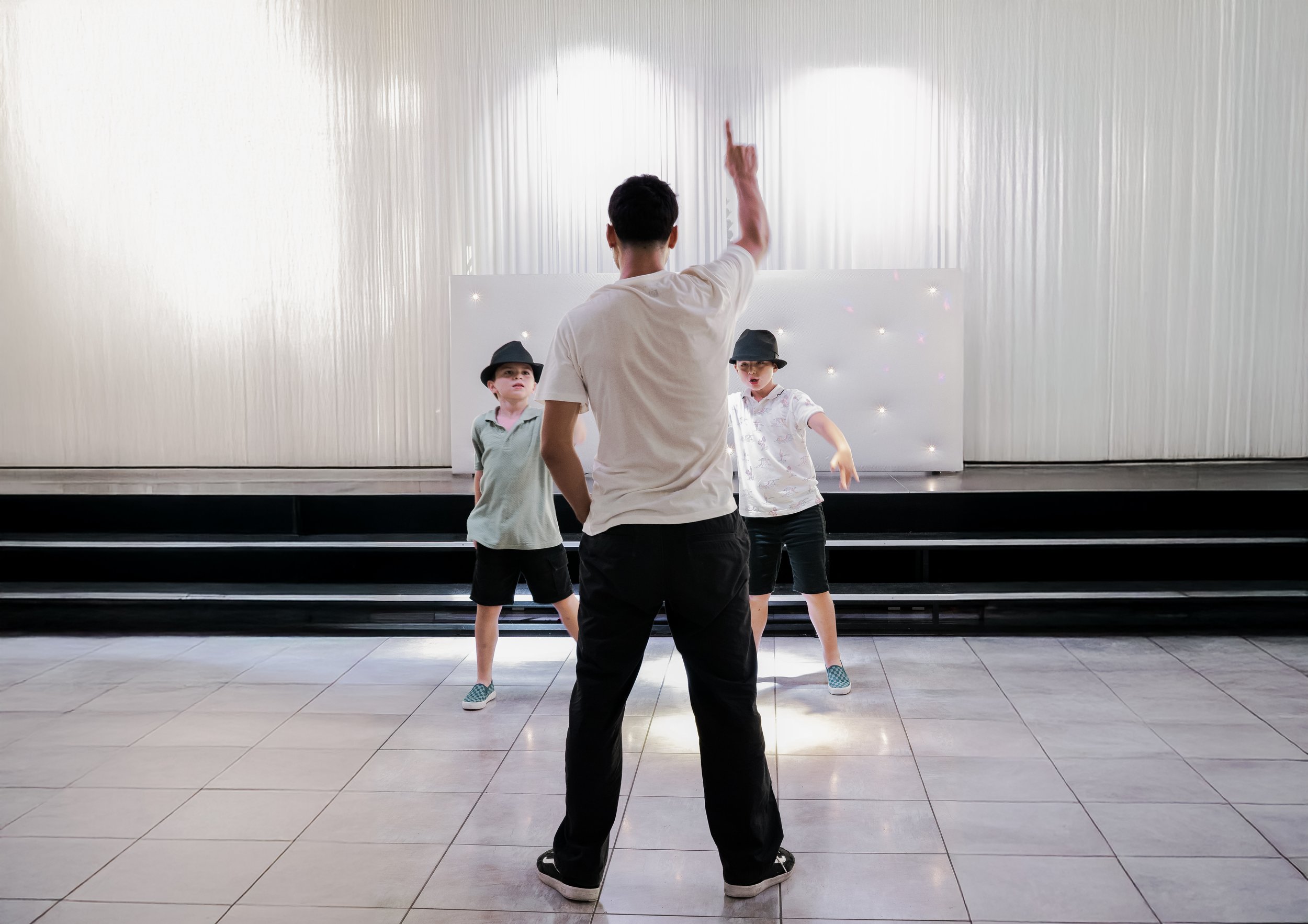 Un jeune homme enseigne à deux enfants comment danser dans une pièce lumineuse, avec une scène au fond.